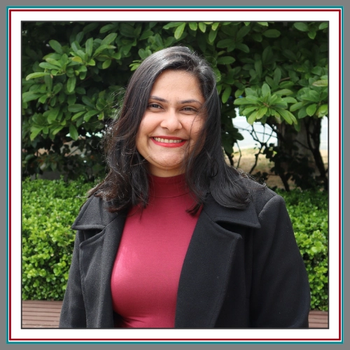 Professional headshot of Anu Singh, team member at Pathway Legal, smiling warmly in a garden setting wearing a crimson top and black blazer.