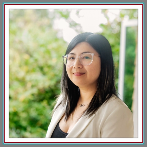 A warm, natural outdoor headshot of Celine Ilano, wearing clear-frame glasses and a cream blazer, photographed in soft natural light against a beautifully blurred green garden backdrop. 