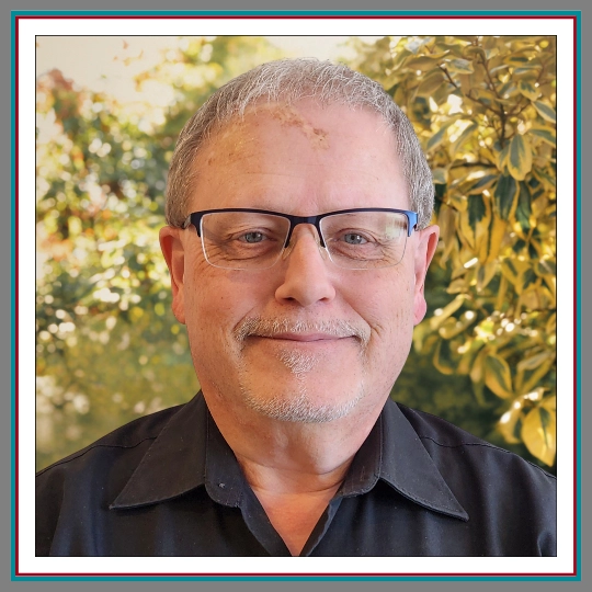 Professional headshot of Curtis, Paralegal at Pathway Legal, smiling warmly with short grey hair, rectangular dark glasses, and a black shirt against a soft green and gold foliage background.