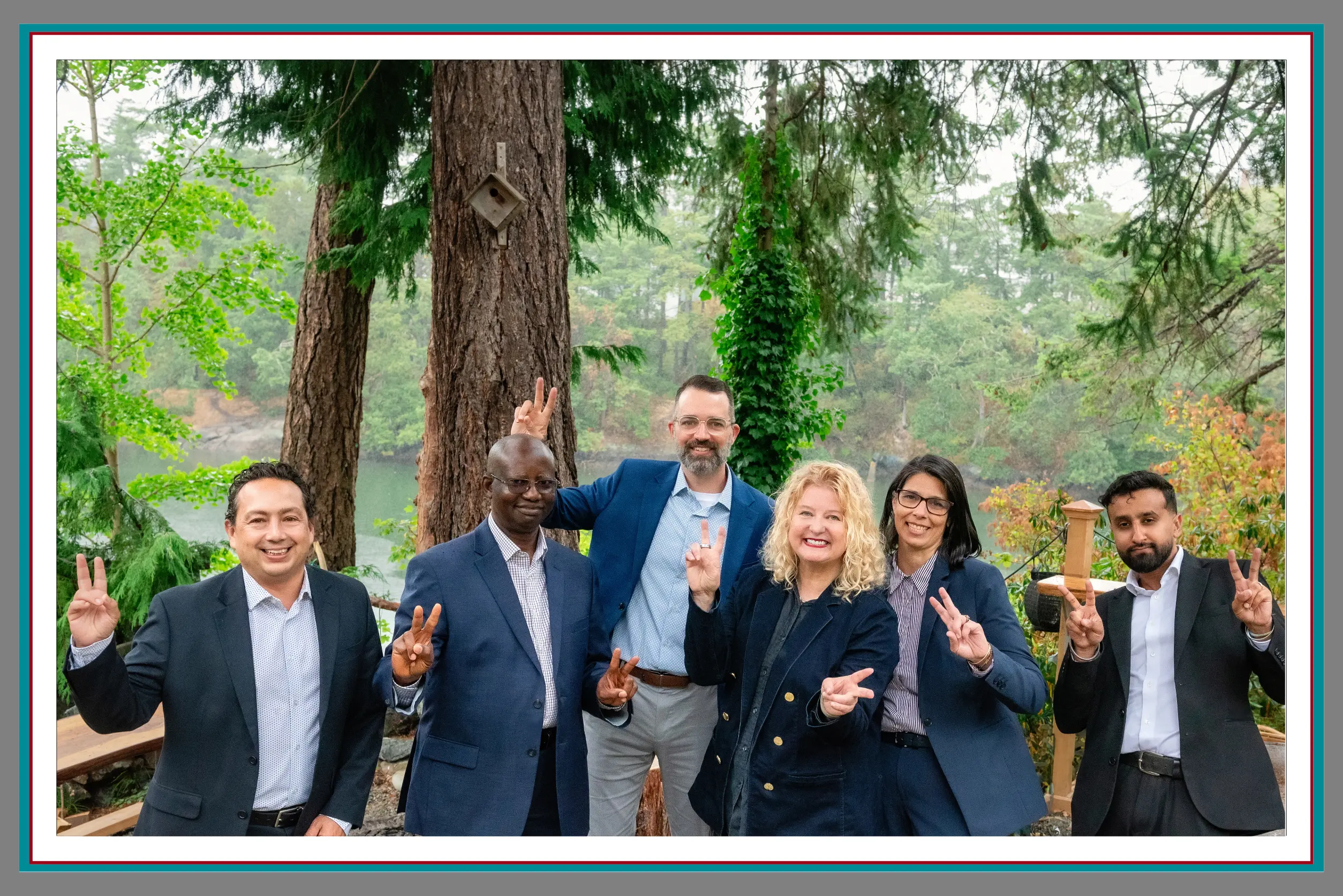 Six lawyers from Pathway Legal standing together outdoors in front of a wooded riverside backdrop, smiling and holding up peace signs. The image reflects their collaborative spirit, approachability, and commitment to supporting families through legal challenges.