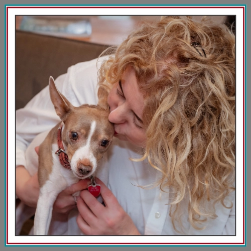 A warm candid close-up of Val Hemminger, founder of Pathway Legal, gently kissing her small fawn and white dog Inu who wears a red heart-shaped tag, captured in a cozy indoor setting.
