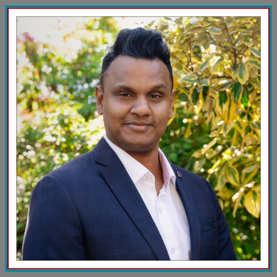 Professional outdoor headshot of Arun Madhavan, family lawyer at Pathway Legal, wearing a navy suit and white shirt, smiling confidently against a warm foliage background.