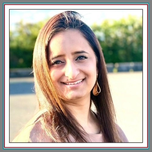 A warm, approachable outdoor headshot of Guneet Kohli, smiling confidently with long straight auburn hair and gold drop earrings, photographed in natural light against a soft blurred outdoor setting.