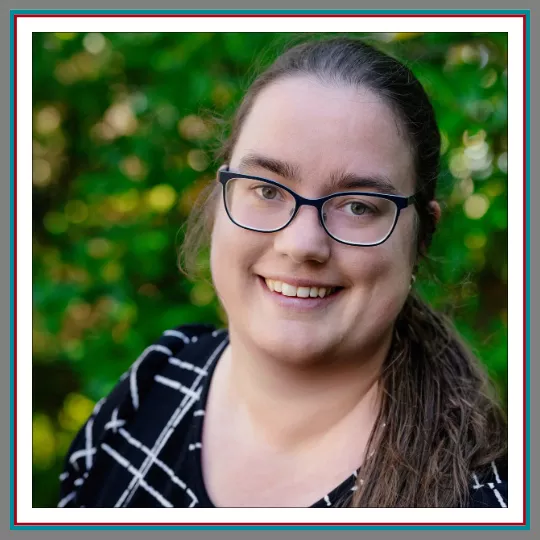 Professional outdoor headshot of Julia Lamoureux, Legal Assistant at Pathway Legal, smiling warmly wearing rectangular dark glasses and a black and white patterned top against a lush green background.