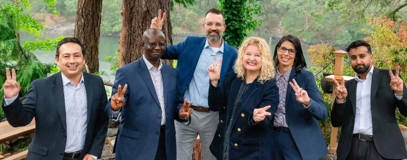 Six lawyers from Pathway Legal standing together outdoors in front of a wooded riverside backdrop, smiling and holding up peace signs. The image reflects their collaborative spirit, approachability, and commitment to supporting families through legal challenges