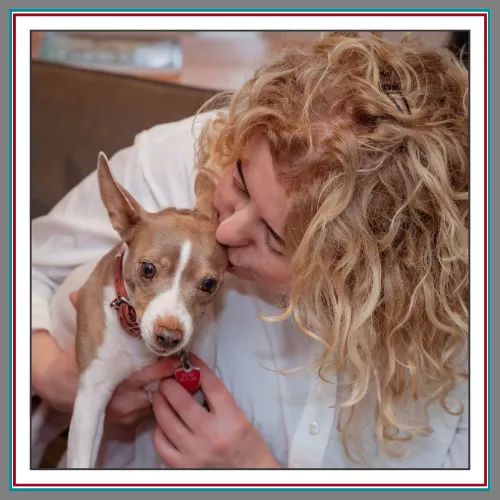 A warm candid close-up of Val Hemminger, founder of Pathway Legal, gently kissing her small fawn and white dog Inu who wears a red heart-shaped tag, captured in a cozy indoor setting.